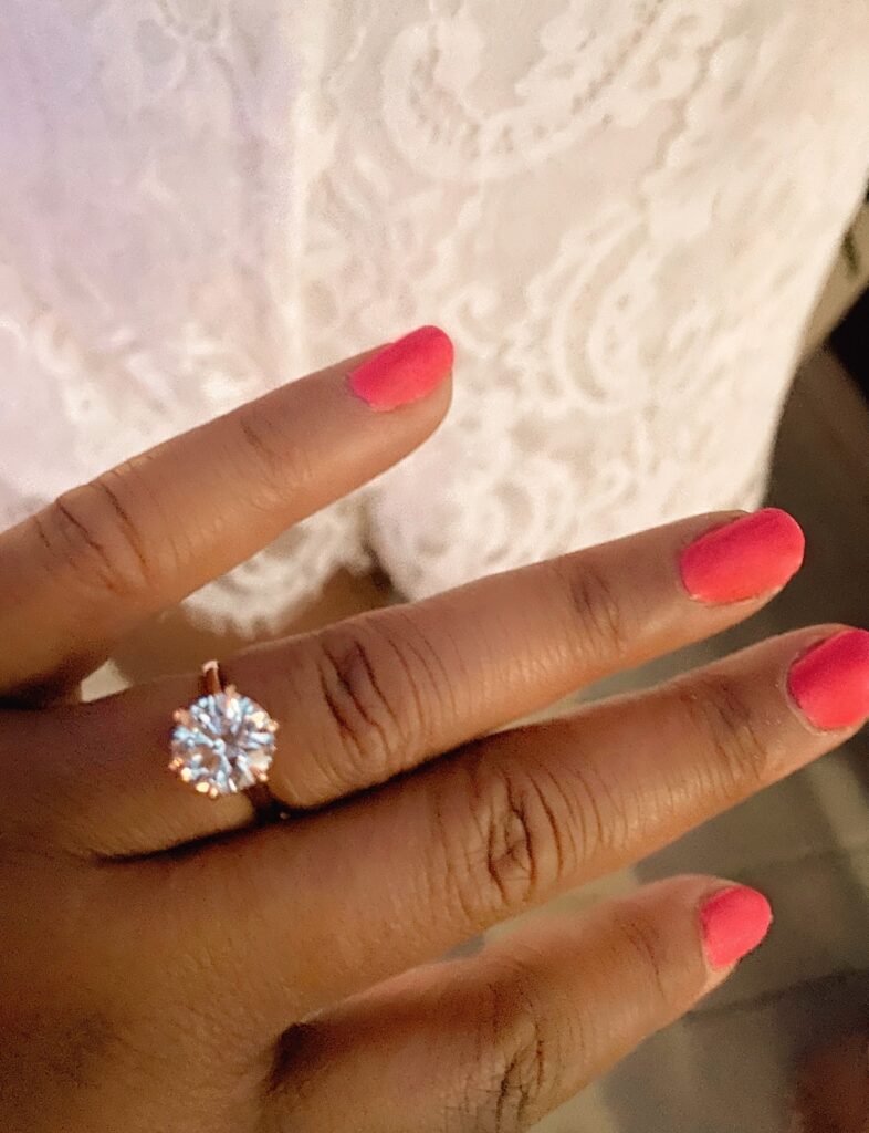 Picture of three carat solitaire diamond engagment ring set in rose gold on african-american hand with bright deep pink nail polish.